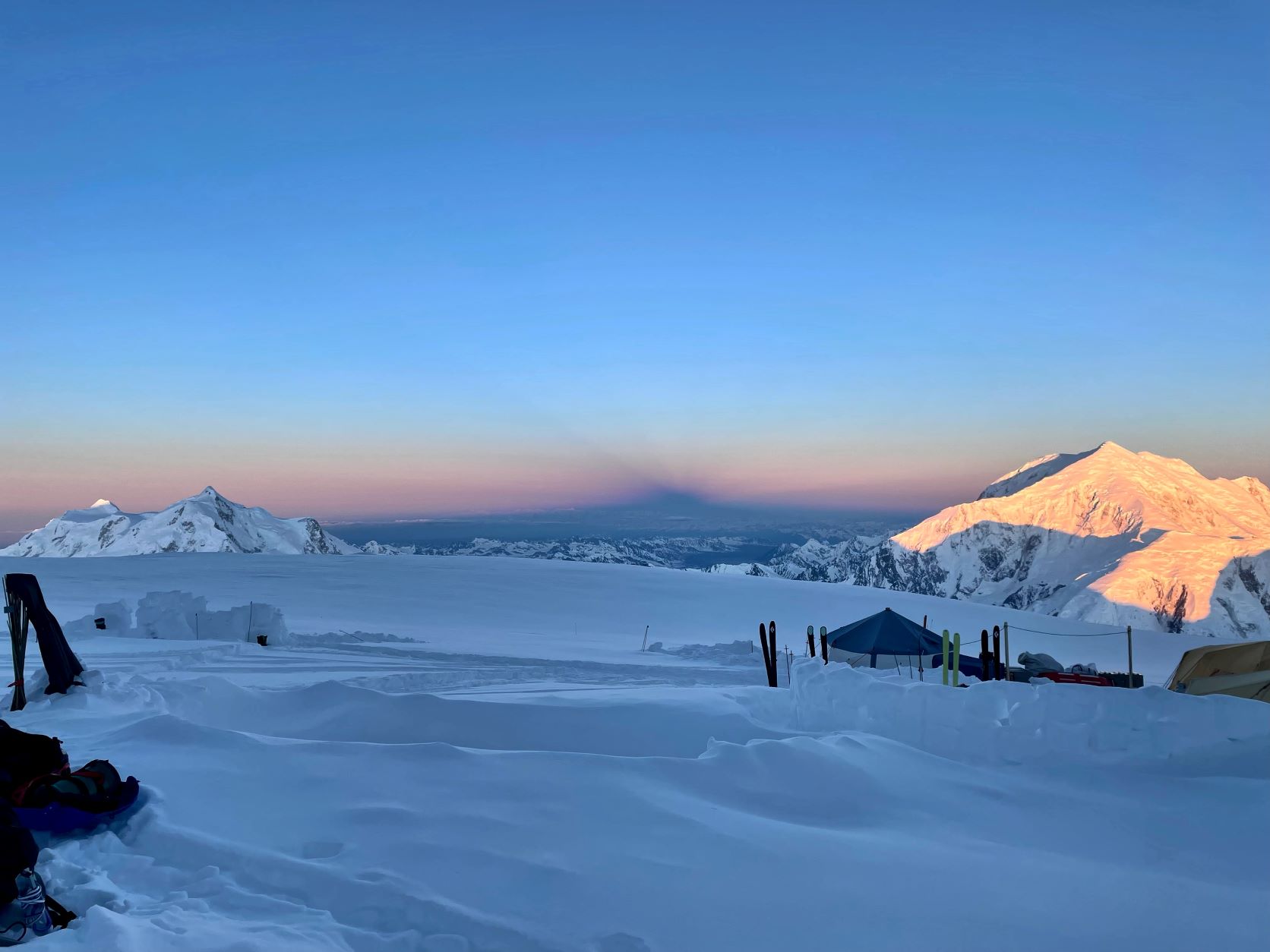 A camp in shadow peers out at an alpenglow peak
