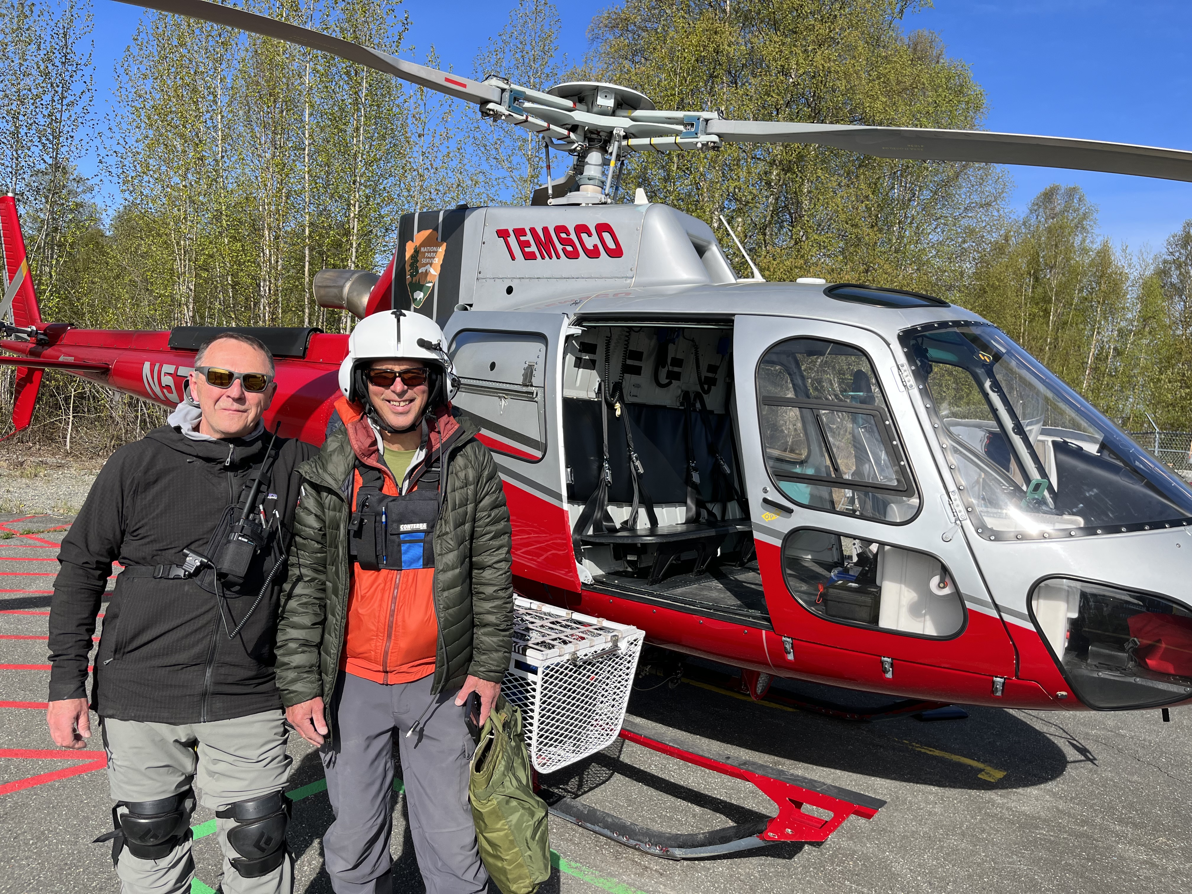 Two mountaineers stand outside the open door of a helicopter on a tree-lined airstrip