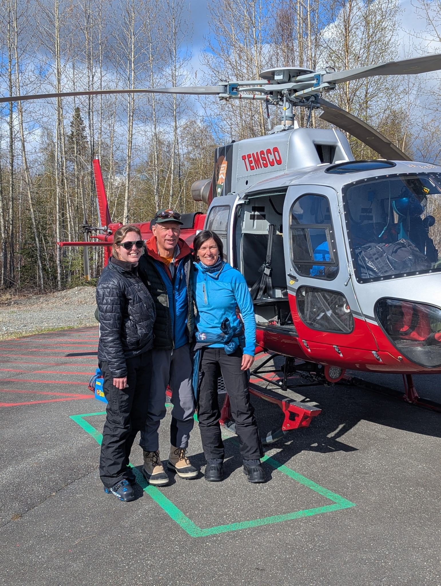 3 people stand in front of a silver and red helicopter