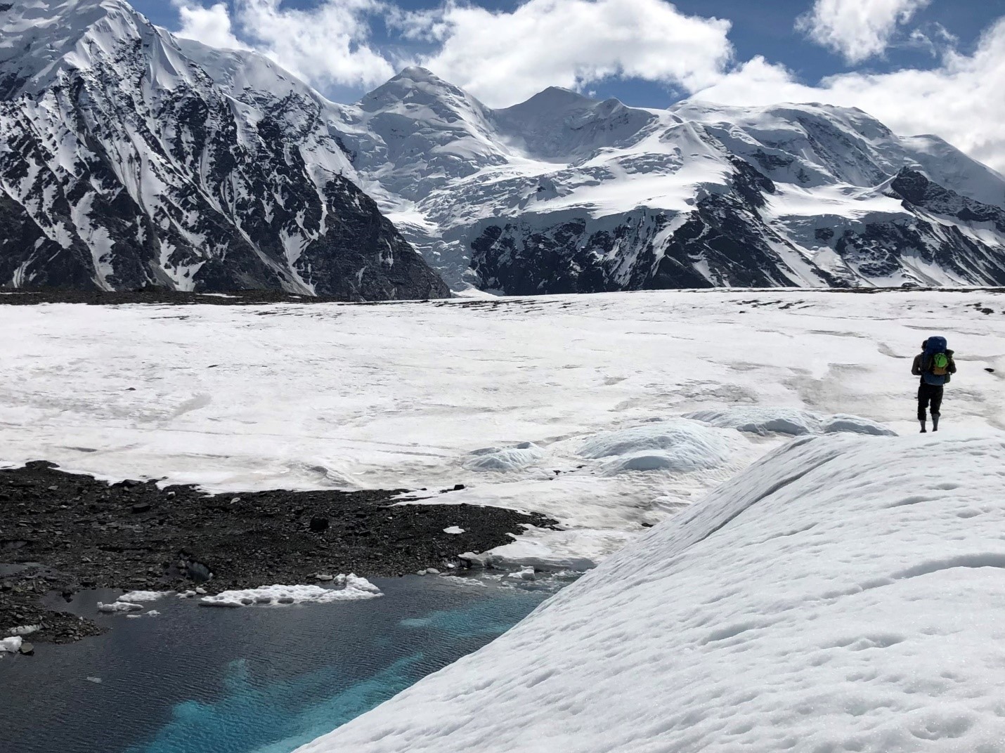 A silhouette of a climber with a backpack hiking on a snowy glacier.  A turquoise blue pool lies to the lower left of the glacier, with rocky peaks in the distance