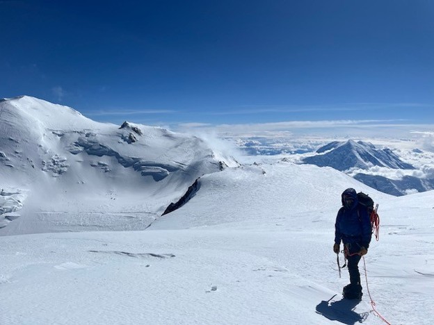 A climber in gear and roped with a red rope walks across a snowy landscape towards mountain peaks and a blue sky.