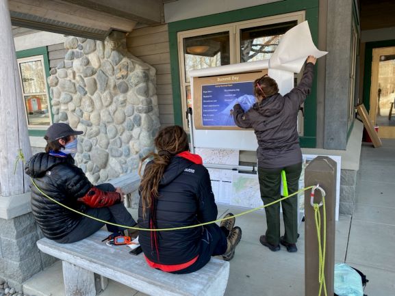 Old-school, open-air, climber orientation Two climbers seated on porch bench listen to a ranger describe mountain features on an outdoor display board