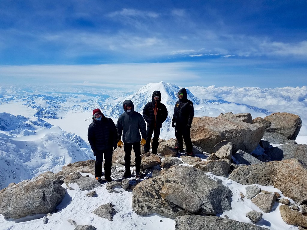 Four well-bundled men stand on a rocky outcropping with high peaks and glaciers in the distance
