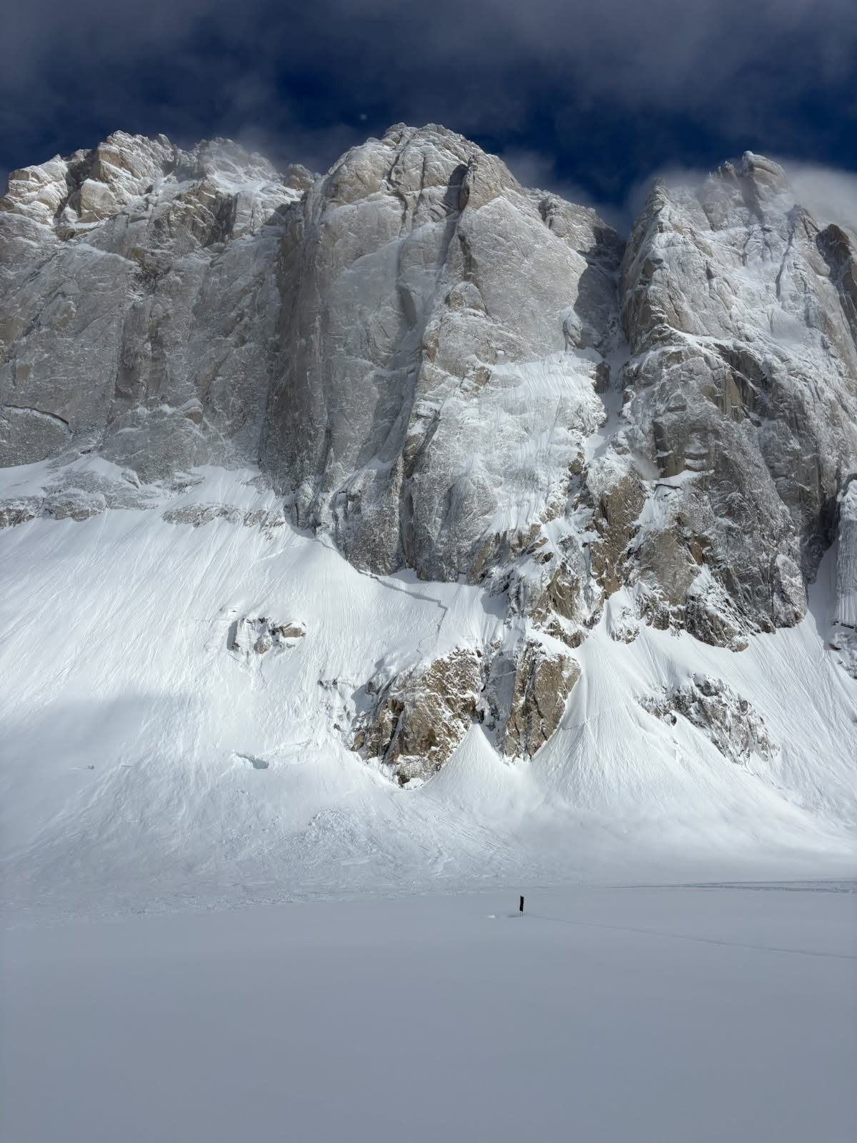 Large white mountain rockface above a snow covered area and a person who looks very small due to the size of the mountain