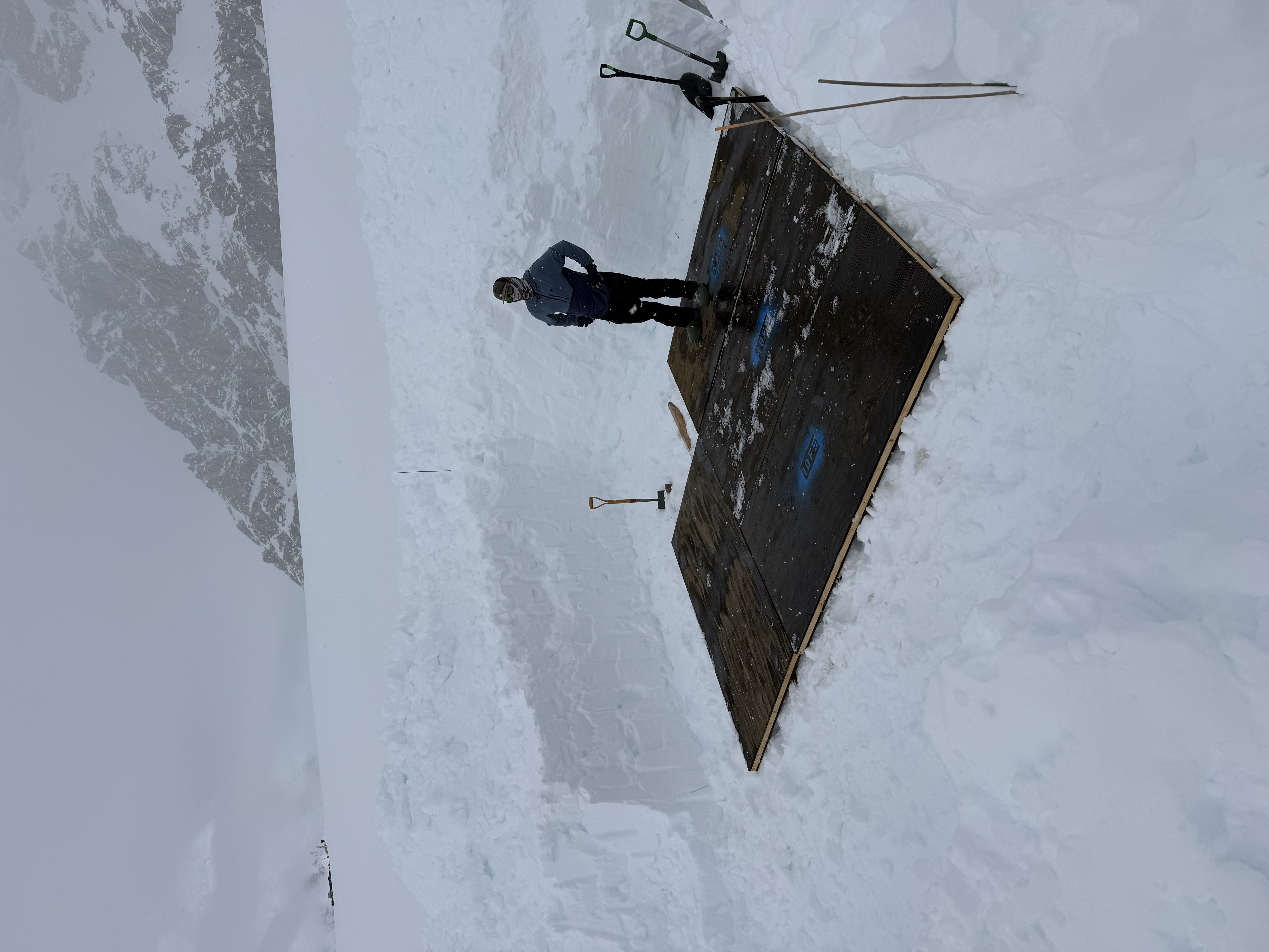A ranger stands in a large square hole dug out of the snow with black flooring covering 