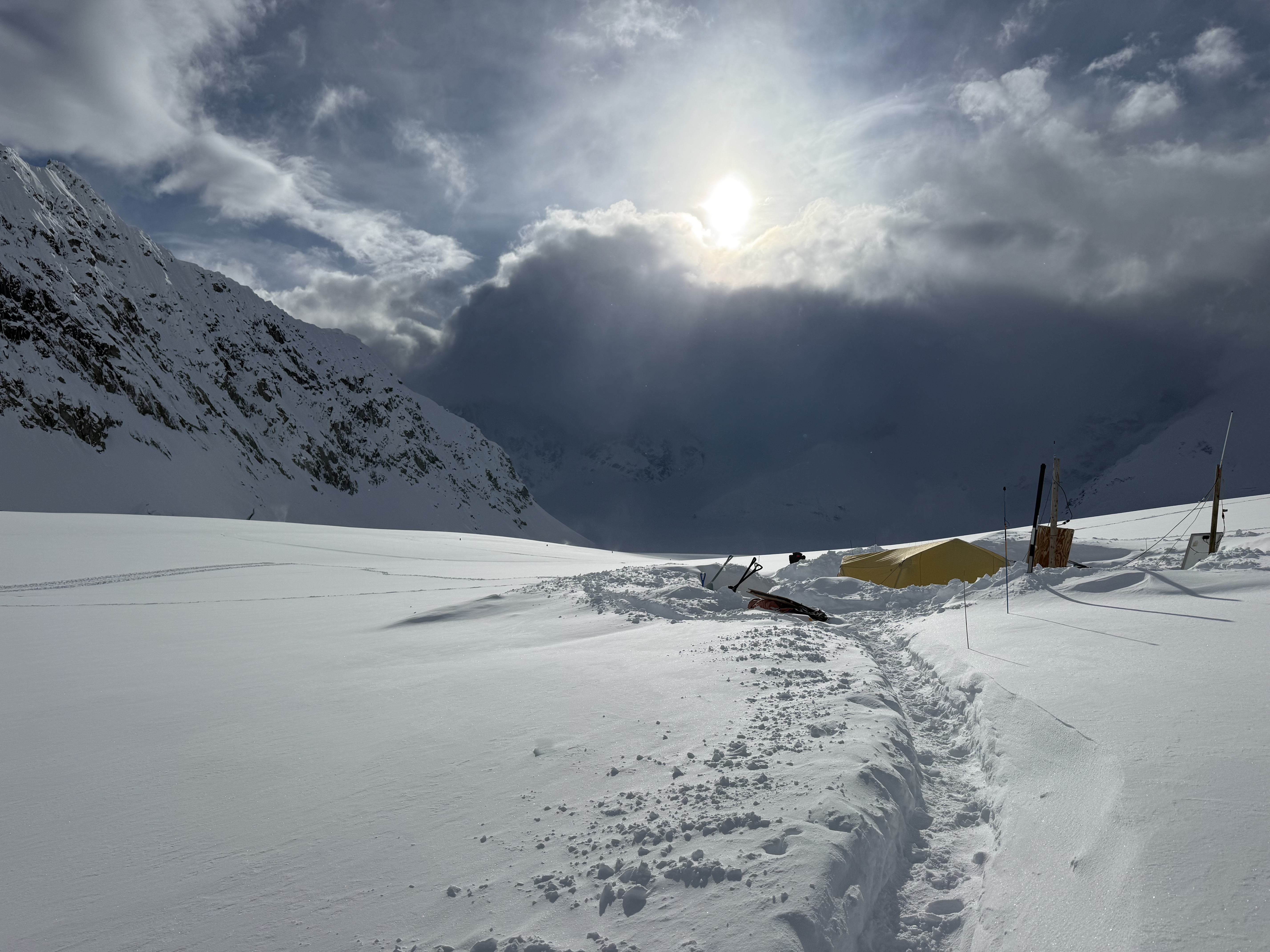 The sun peeks above a large group of grey clouds over a snowy landscape with a trail dug