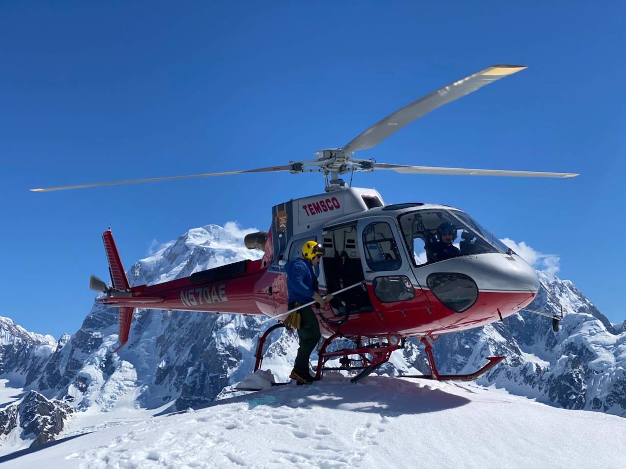 Joe works on the radio repeater Ranger stands outside a helicopter balanced atop a mountain peak