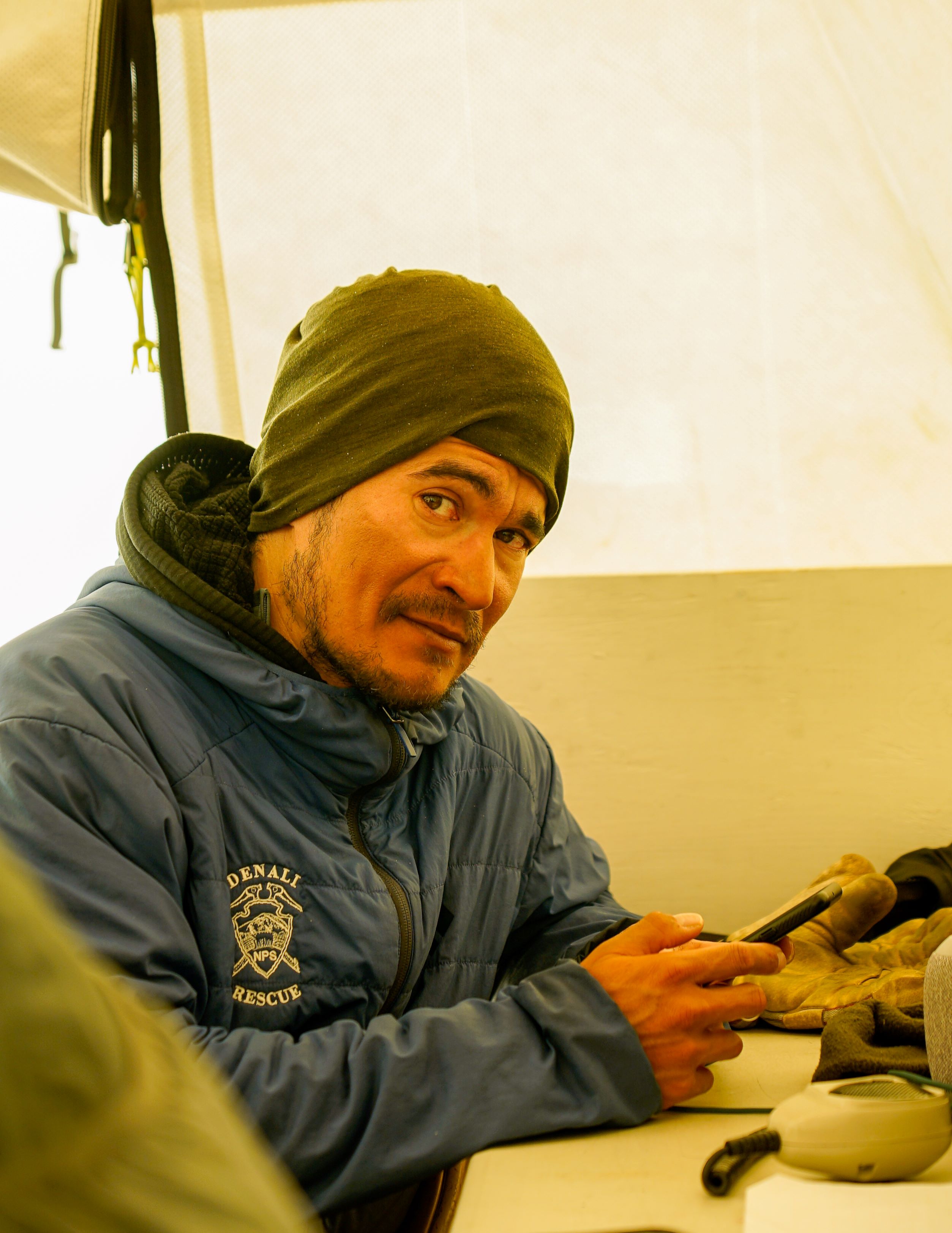 A portrait of a man in a tent on his phone wearing a blue jacket that says Denali Rescue