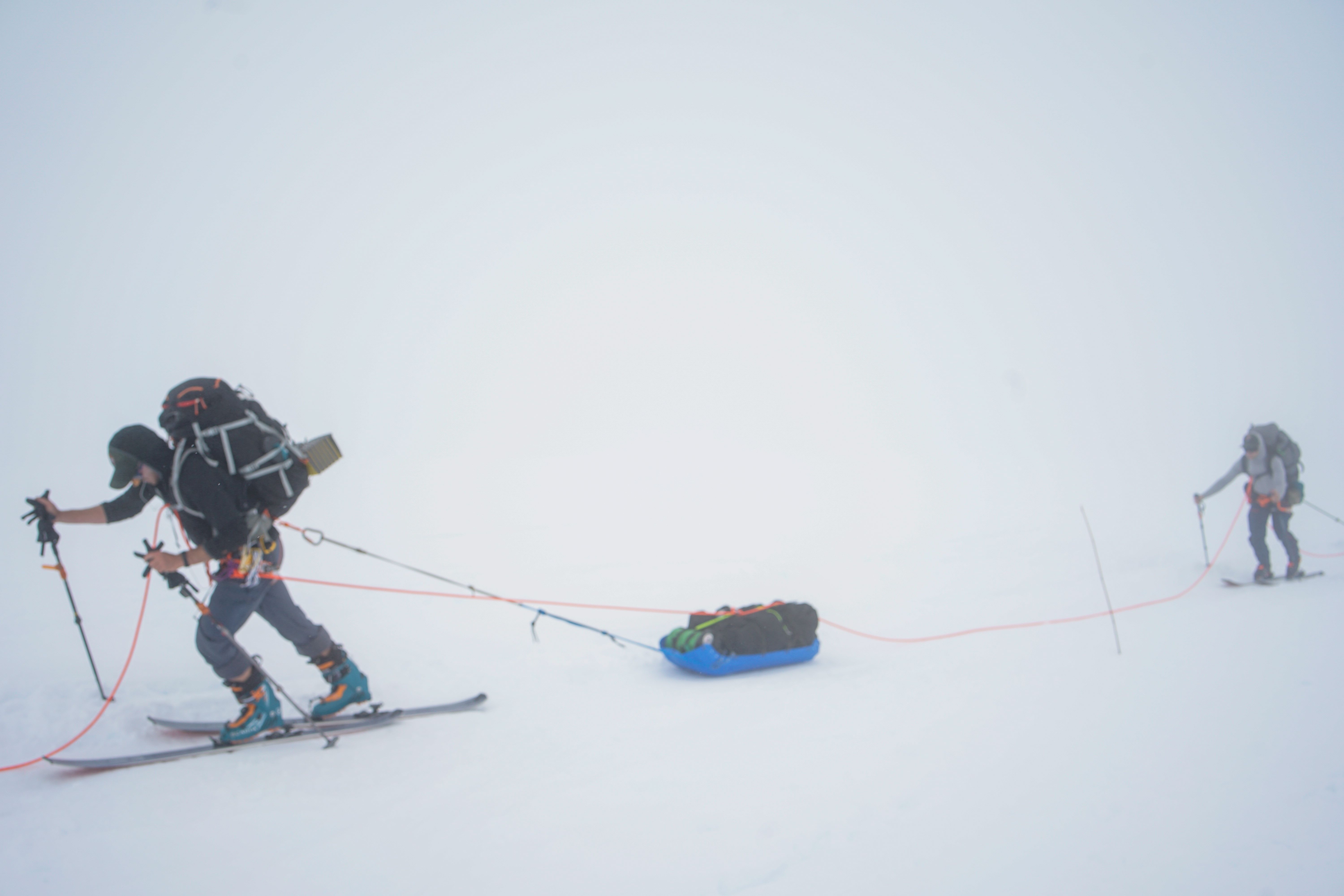 Two roped climbers ski uphill in a whiteout, the lead climber pulling a heavy sled