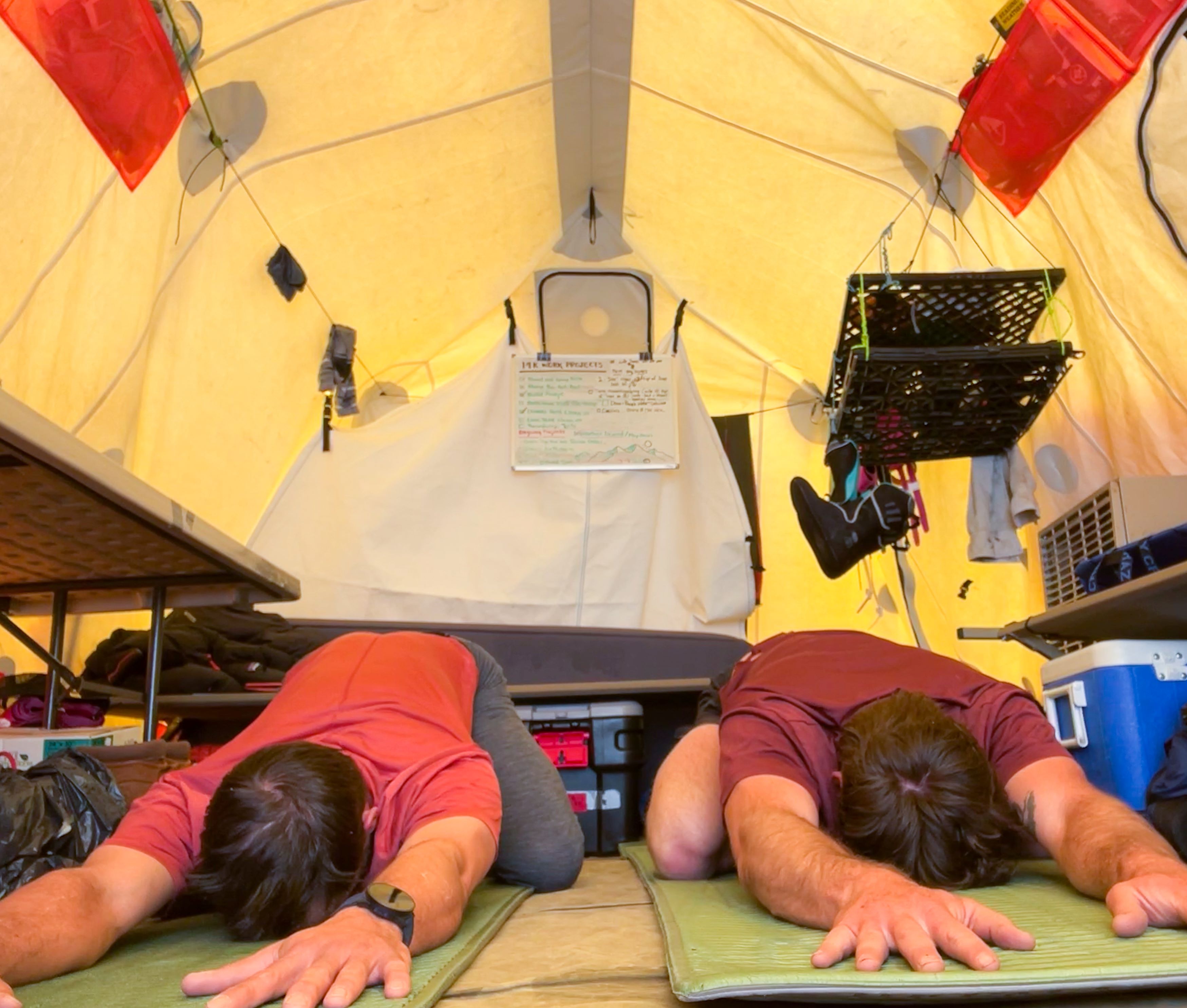 Two men do a yoga stretch on the floor of a yellow tent.