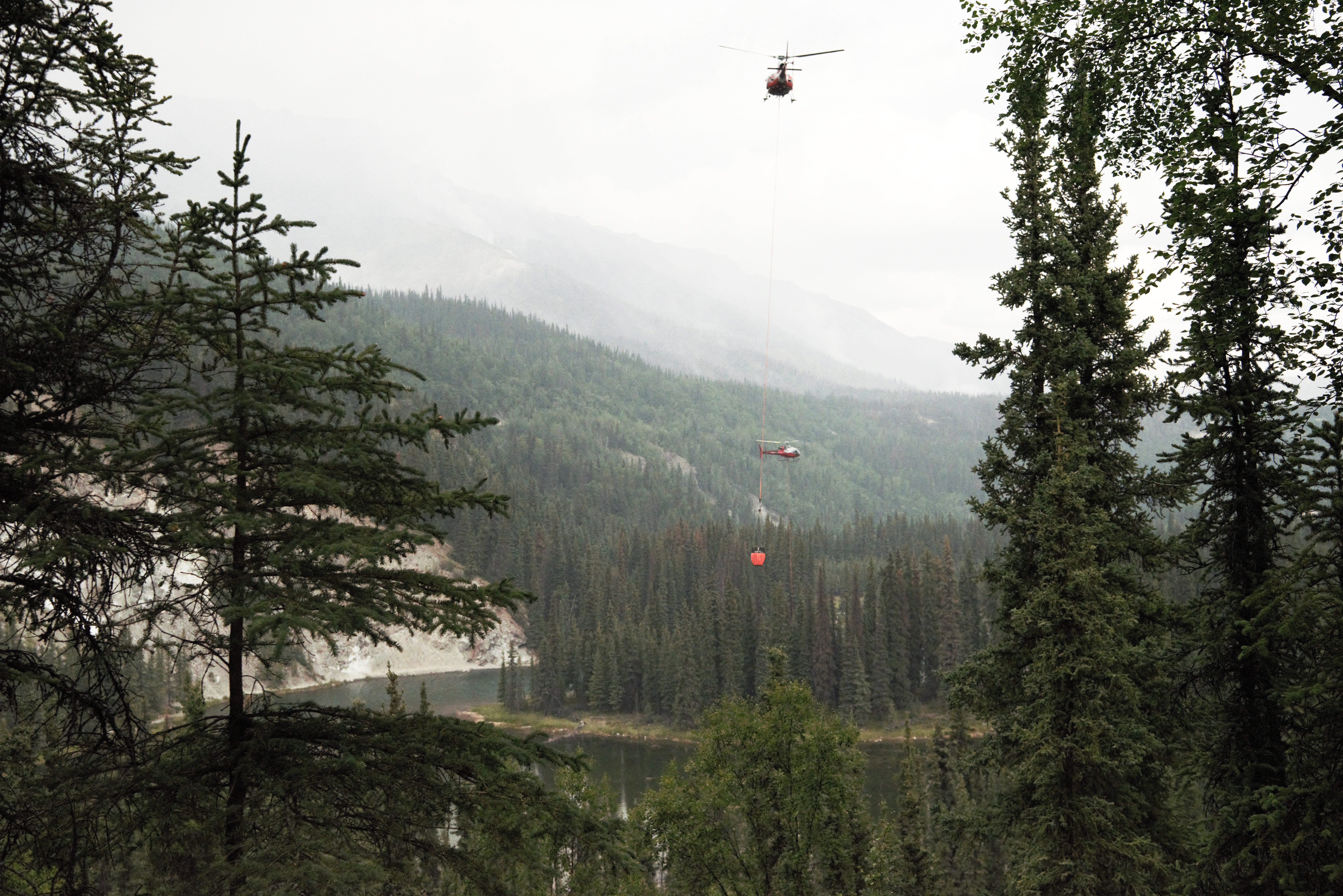 Riley Fire Helos_NPS Photo-E.Miller Two red helicopters drawing water from a lake below using orange buckets. Smoke is rising in the mountainous background