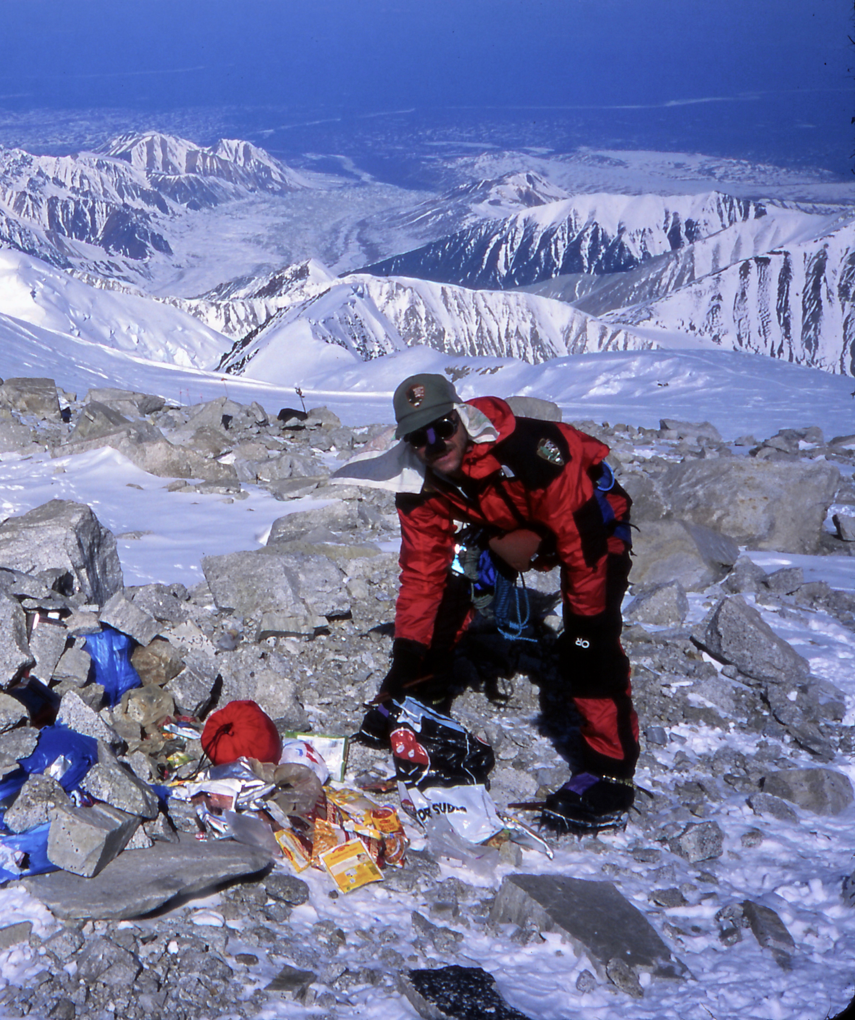 Ranger wearing crampons and mountaineering gear cleans up a pile of trash in a boulder zone on a snowy slope