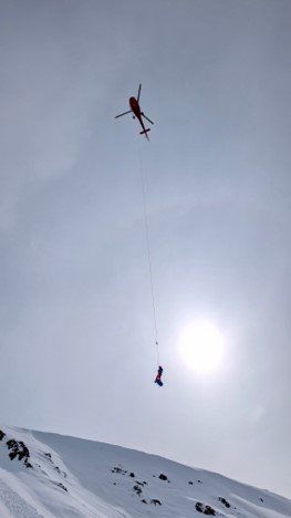 A helicopter dangles equipment above a snowy landscape against a grey sky