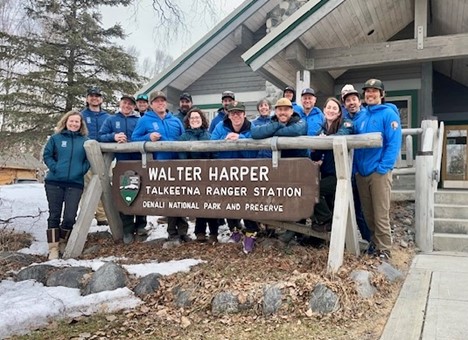 Eleven park rangers in blue jackets stand behind a sign that says Walter Harper Talkeetna Ranger Station Denali National Park and Preserve