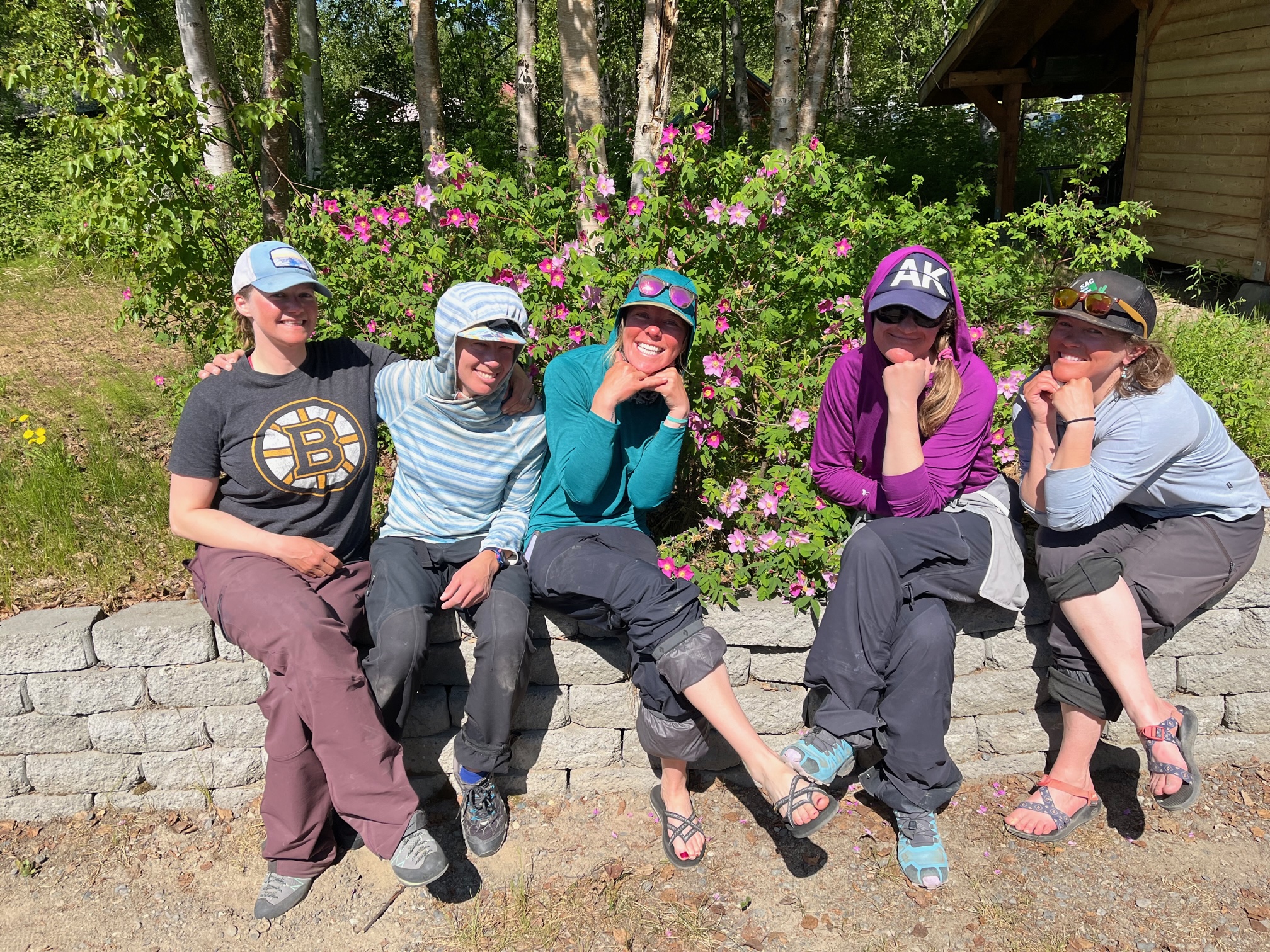The Five women sit on a rock wall in front of a rose bush