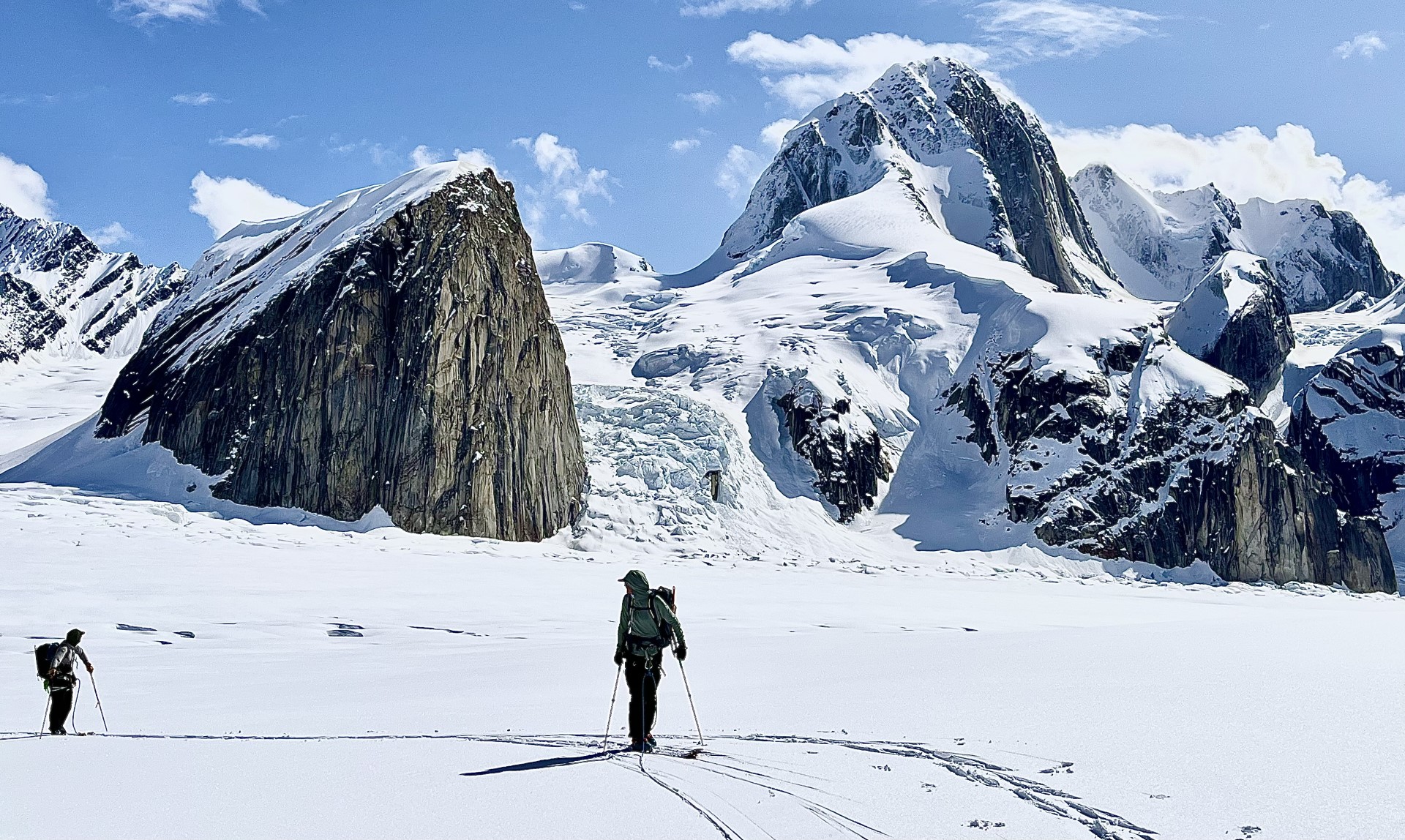 Two skiers in the foreground, with dramatic snow-covered peaks in the backdrop