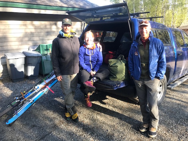 Three mountaineers pose outside a truck with their skis and gear