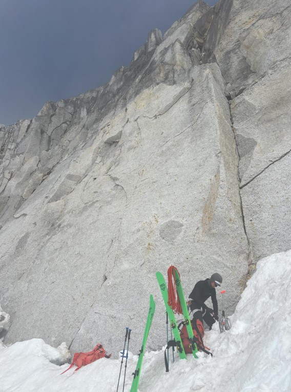 Transitioning at the base of Masters of the Mid on the Crown Jewel A ranger stands next to a rock face with two pairs of green skis