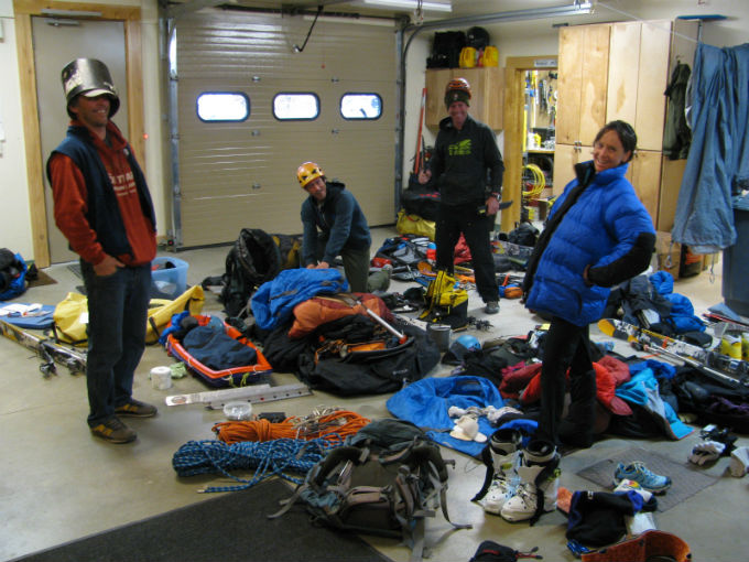 Four climbers stand in a garage amidst a floor full of assorted mountain gear
