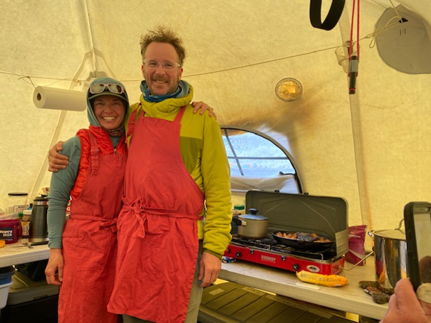Smiling VIPs in matching red aprons prepare dinner on a camp stove