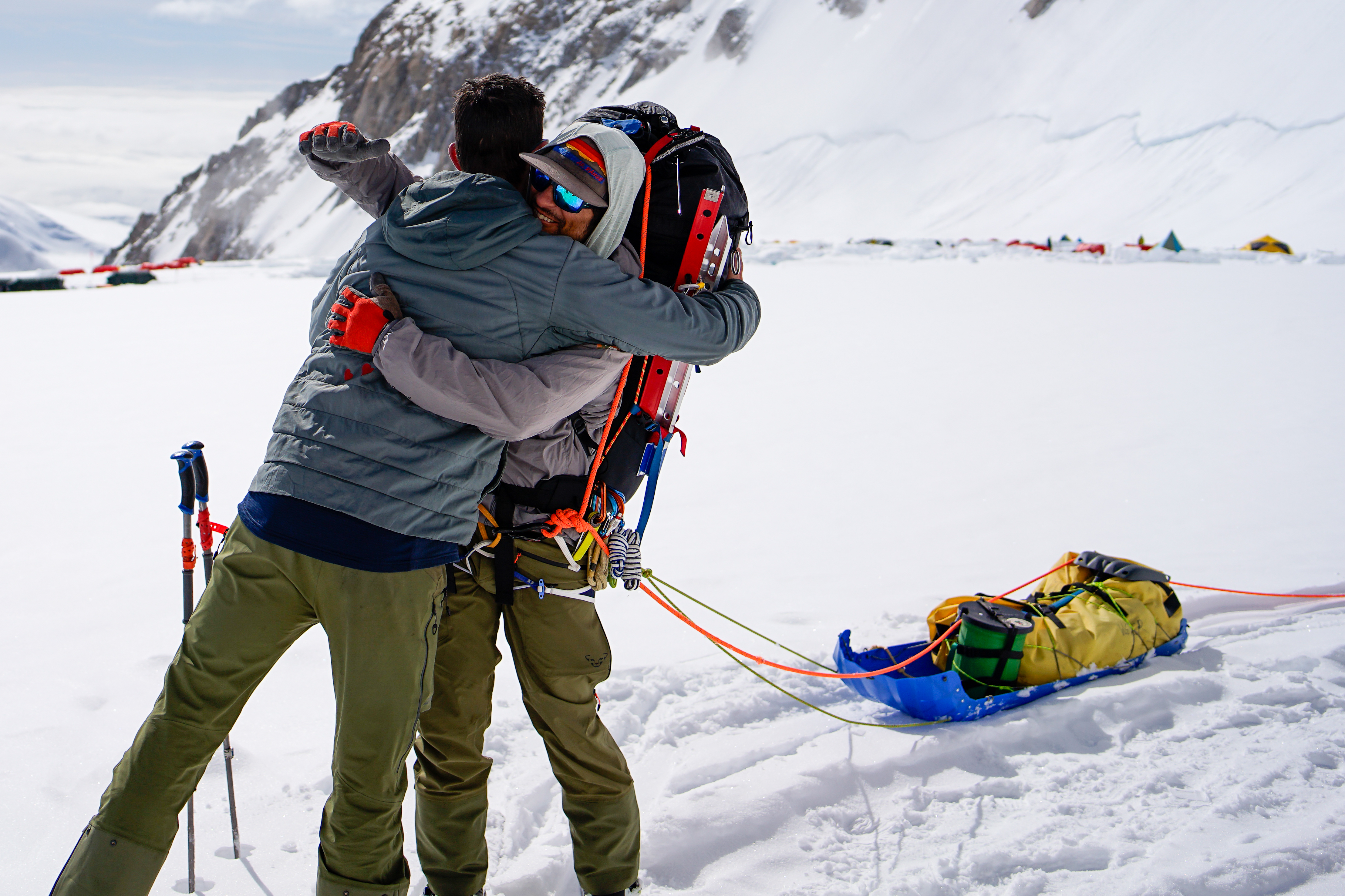 Two climbers embrace, one wearing a huge backpack and pulling a sled. 