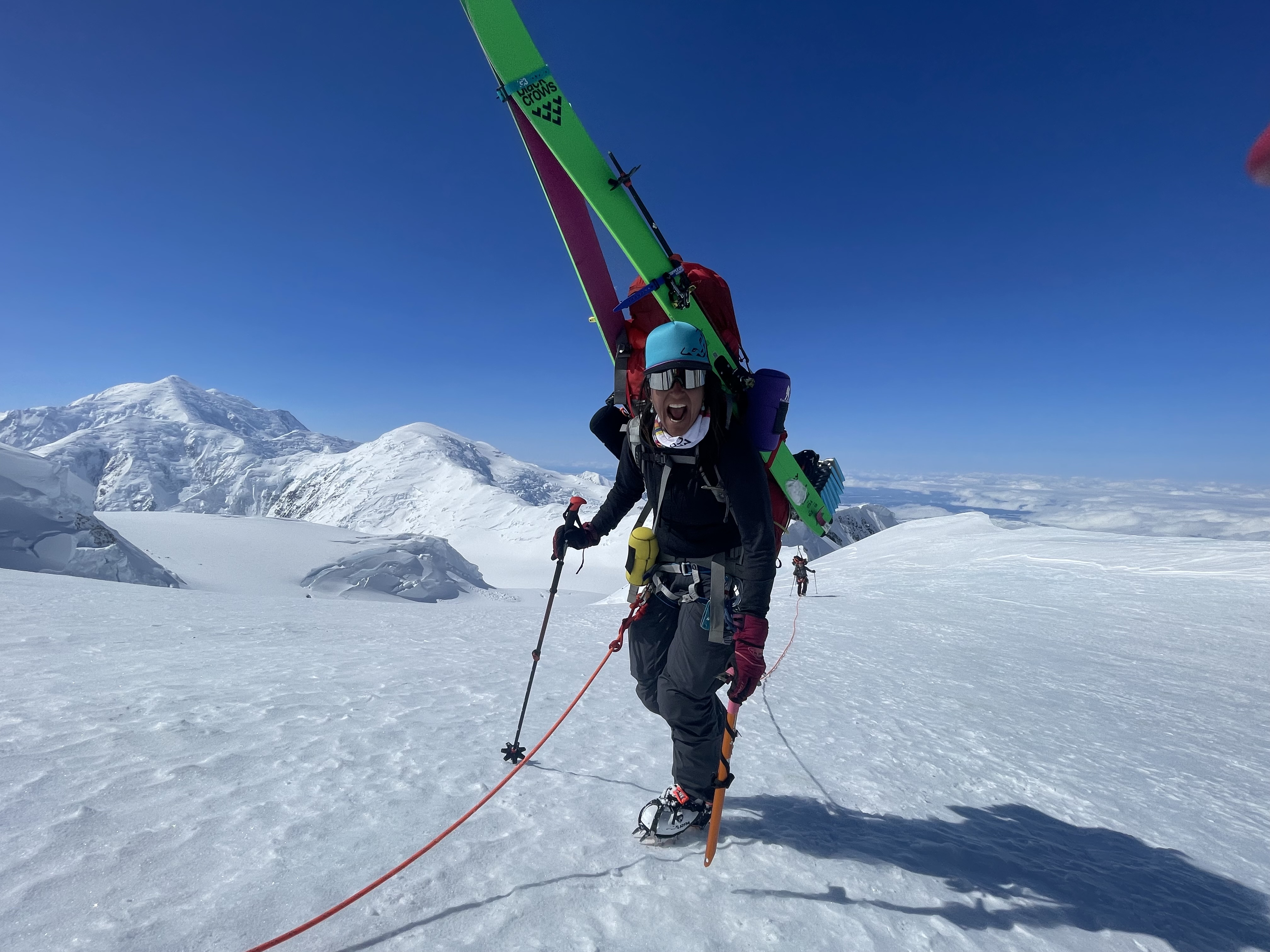 A woman with a huge smile climbs up a mountain slope carrying a huge backback with skis on it