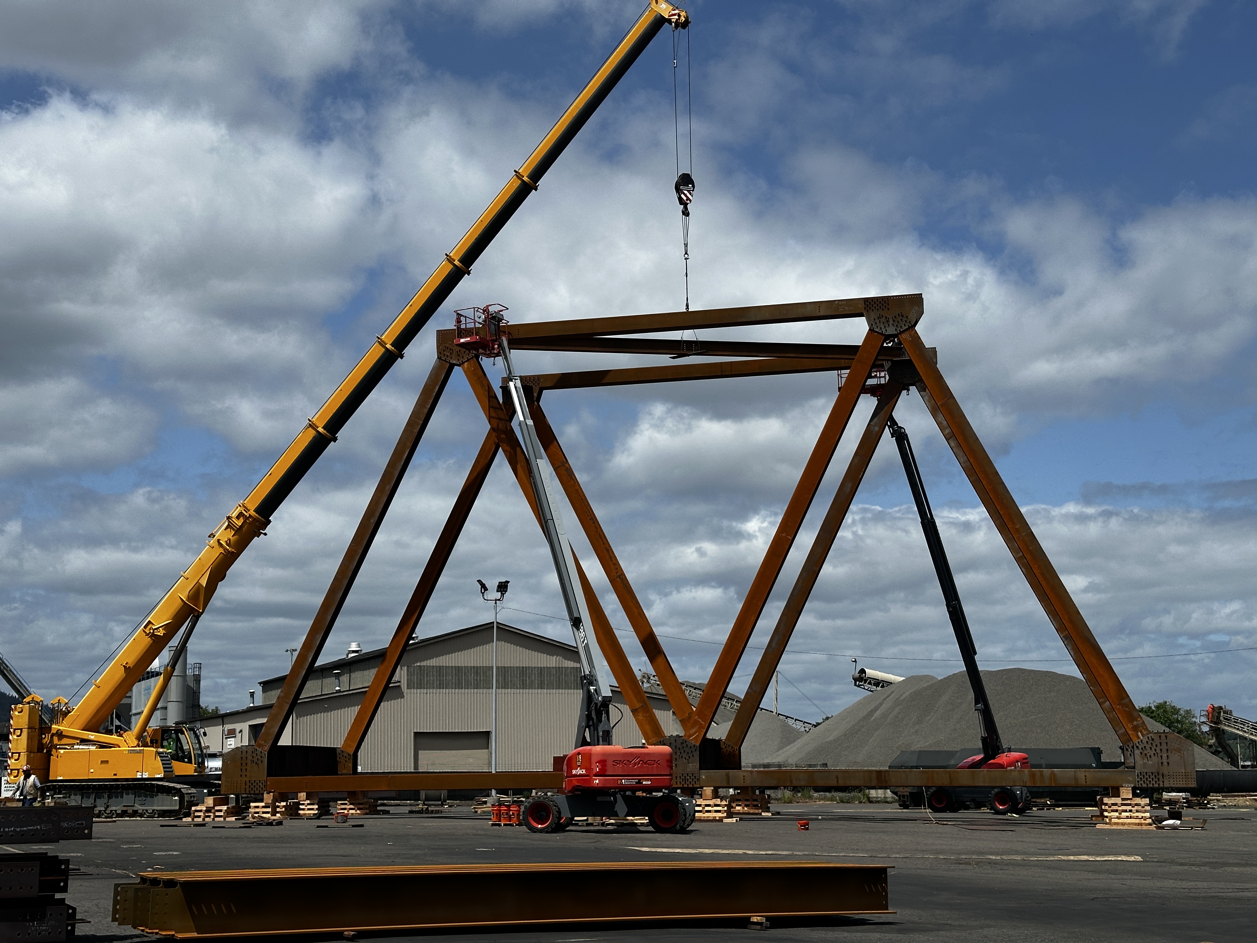 bridge trial assembly In a construction yard, several large steel beams have been attached to create part of a bridge truss. A crane lowers another piece into place.