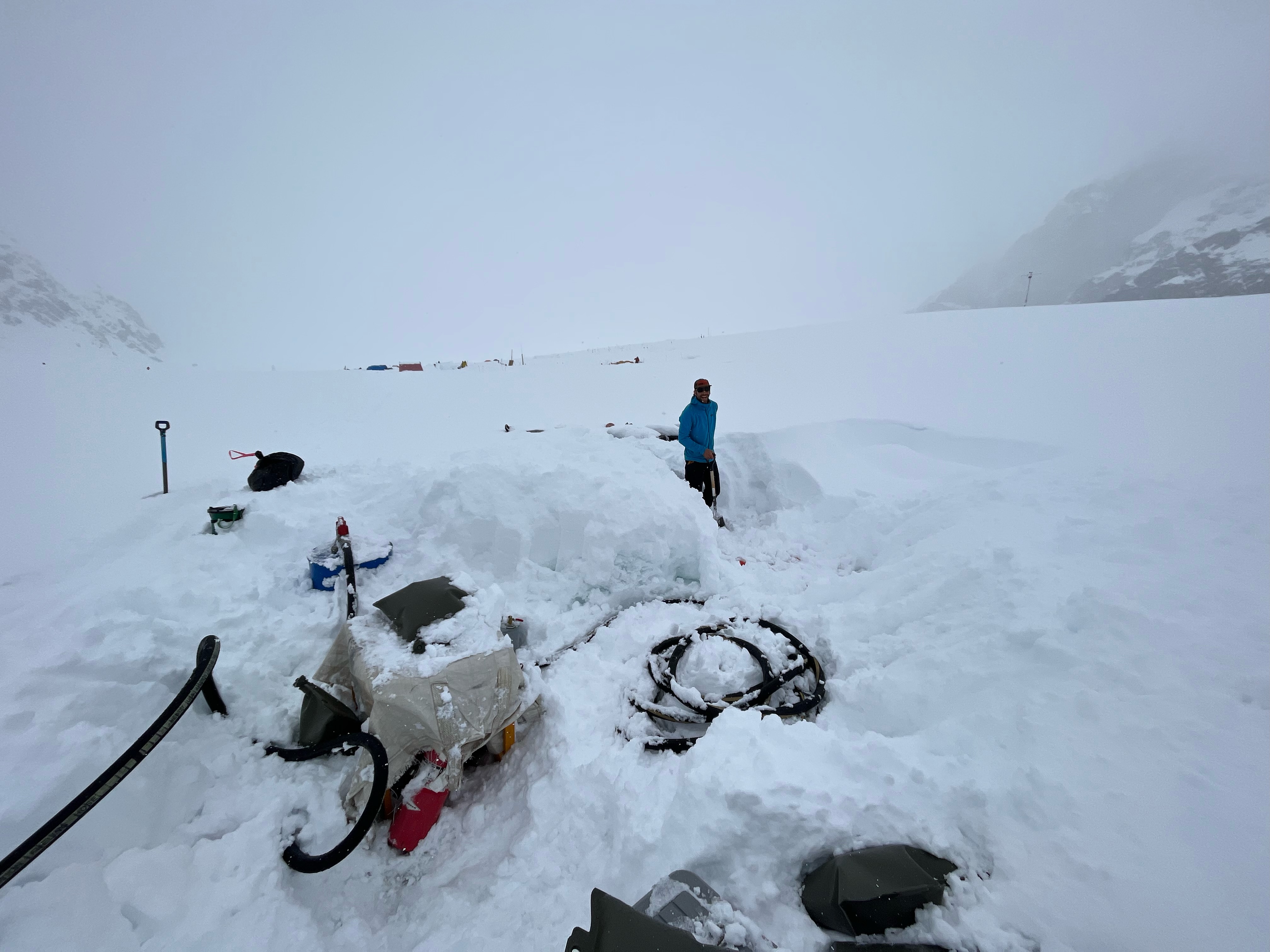 Partially snow-covered fueling equipment sits on a glacier while a man with a shovel looks on