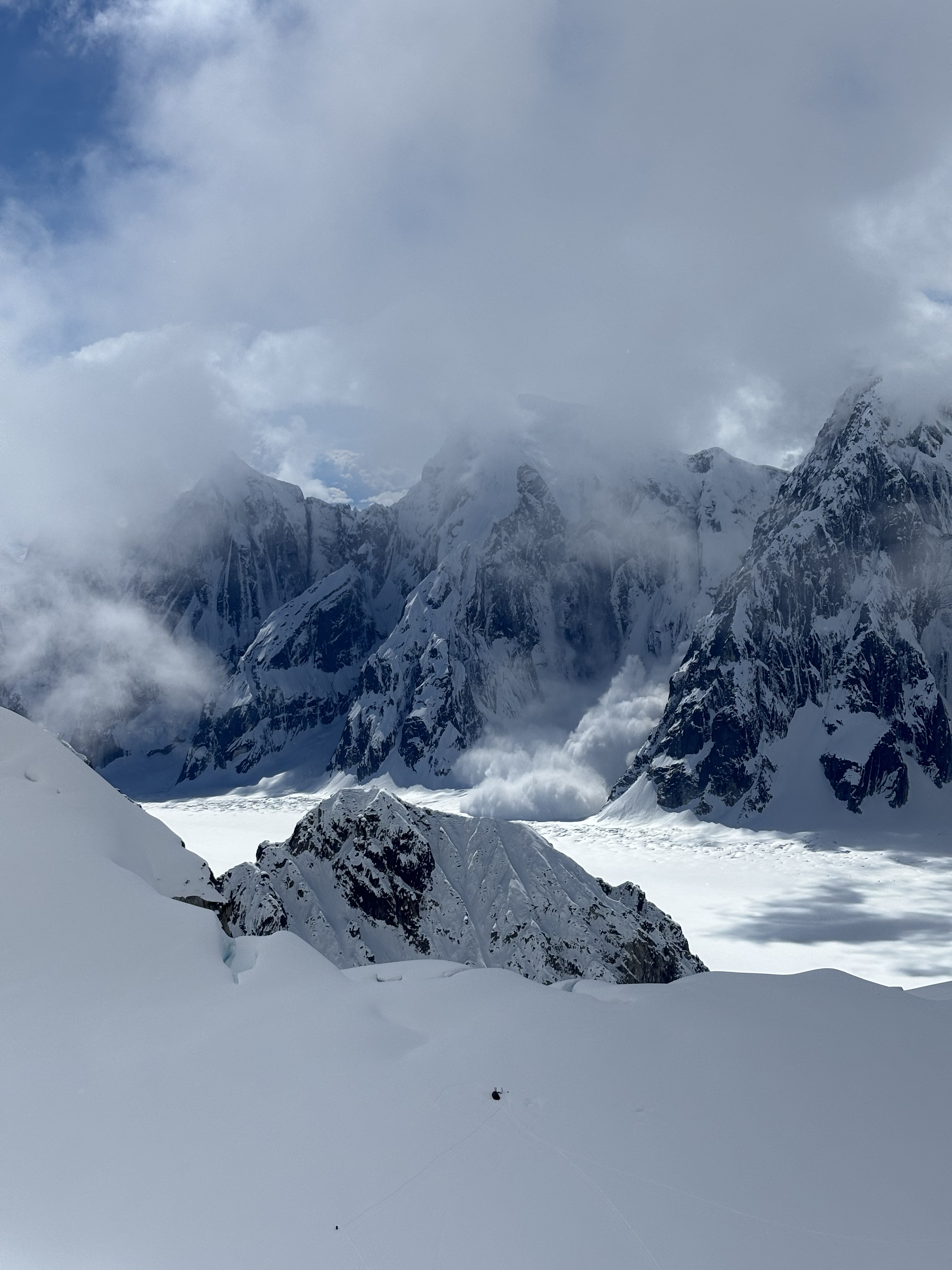 large serac-icefall avalanche Grey snowy peaks with a wall of snow sliding down to a snow-covered field with a large group of clouds above.