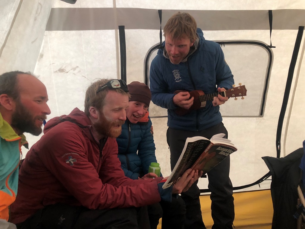 Band Camp Four climbers in a tent sign from a music book as one plays the ukelele