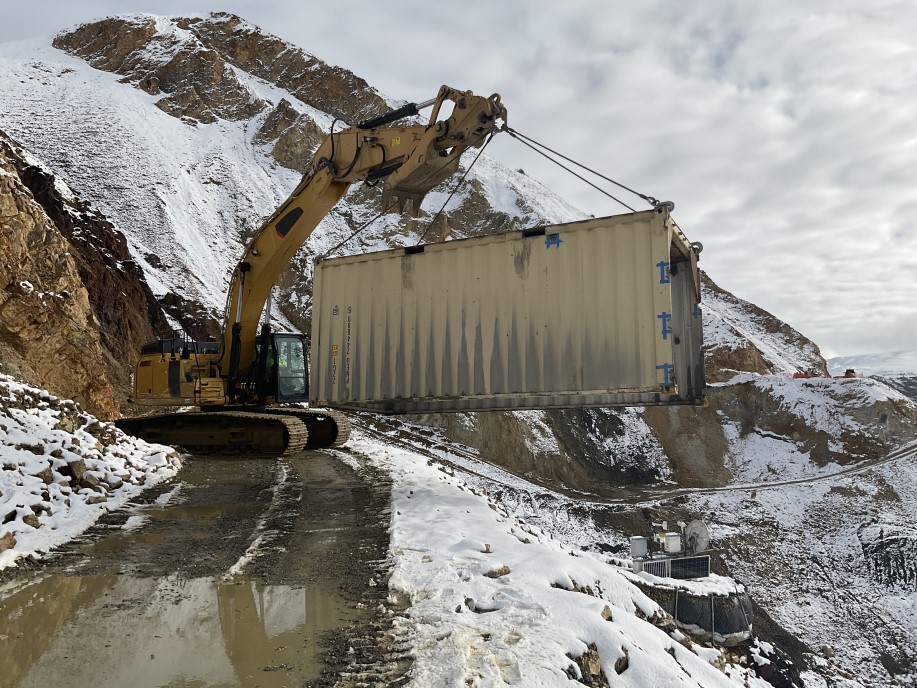 Large machinery lifts a cargo container and moves it over the edge of the road towards the pretty rocks monitoring station instruments on the slope below.