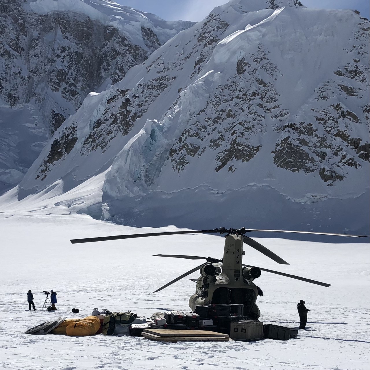 Chinook military helicopter sits on a glacier at the base of a mountain slope, surrounded by a large pile of plastic packing boxes and plywood sheets.