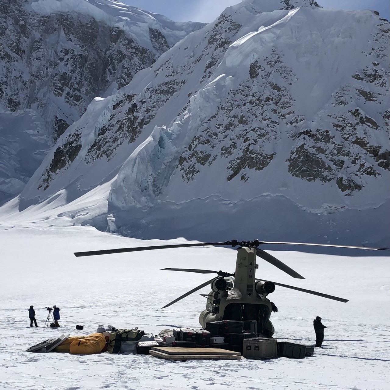 US Army Chinook helicopter surrounded by unloaded camp infrastructure Chinook military helicopter sits on a glacier at the base of a mountain slope, surrounded by a large pile of plastic packing boxes and plywood sheets.