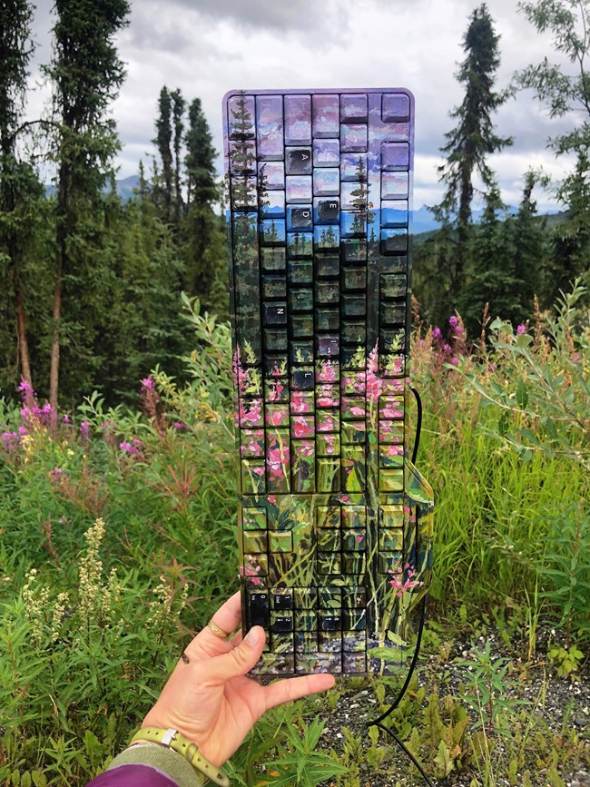 a keyboard painted with flowers and trees, held up against a background of real trees and flowers