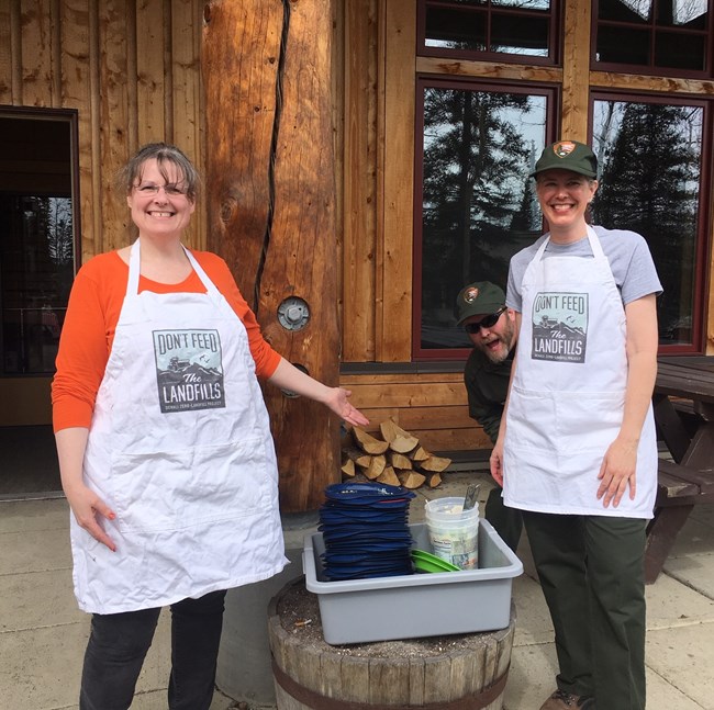Two people wearing aprons that read "Don't Feed the Landfills" smile while standing next to a bucket full of dirty dishes.