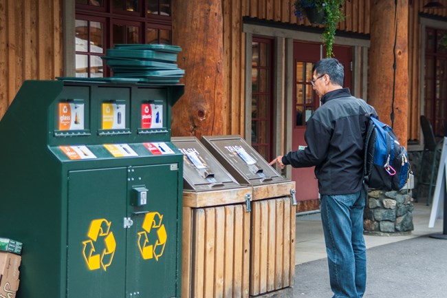 A person looks at a trash receptacle next to three recycling bins.