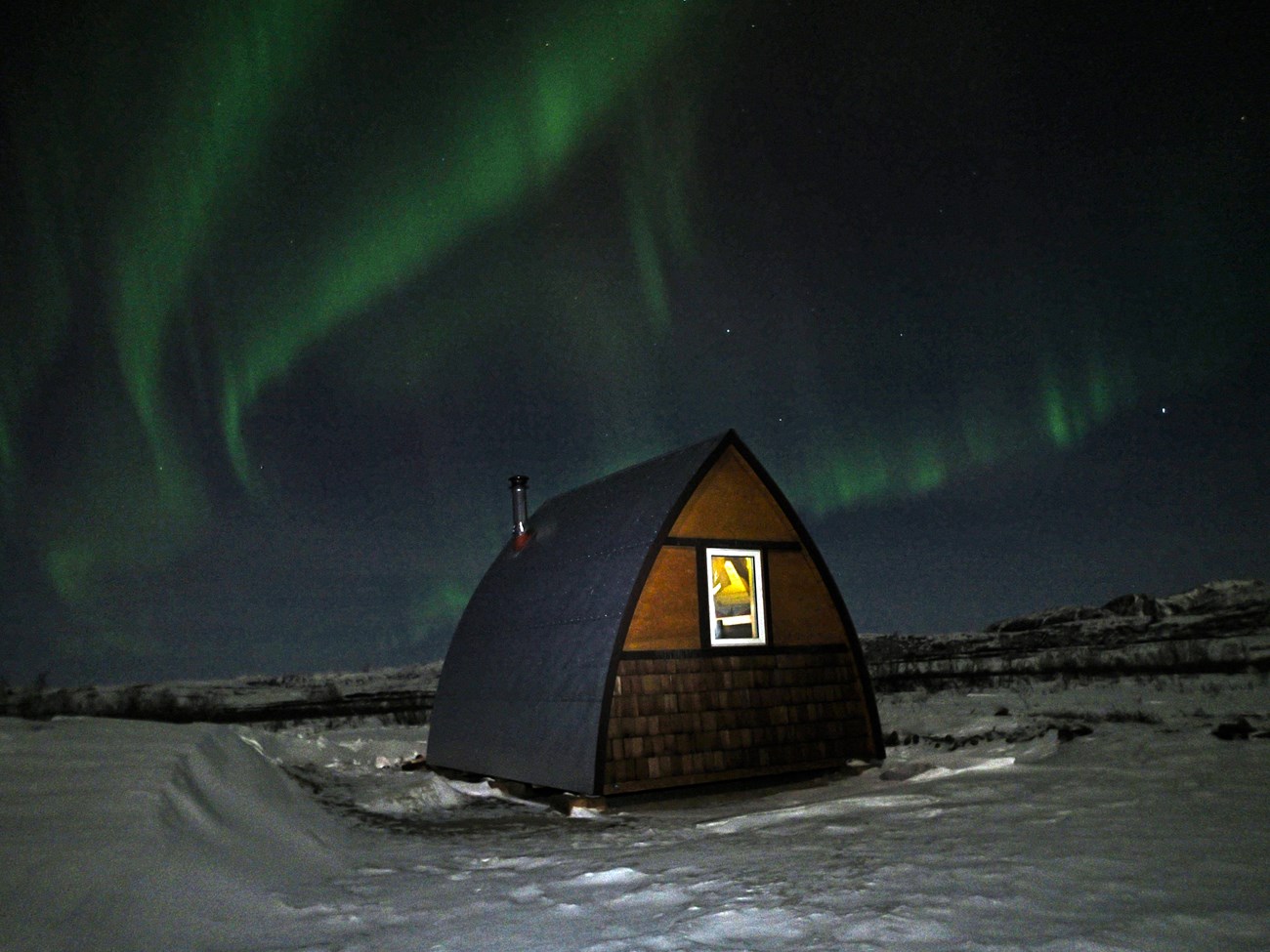 a snowy forest at night with a log cabin with interior lights glowing under a sky full of green aurora
