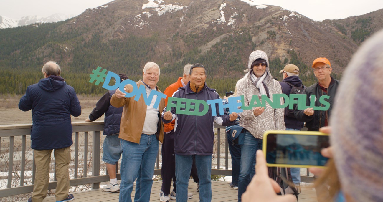 Three people stand on a deck overlooking a river holding a sign that reads Don't Feed The Landfills.