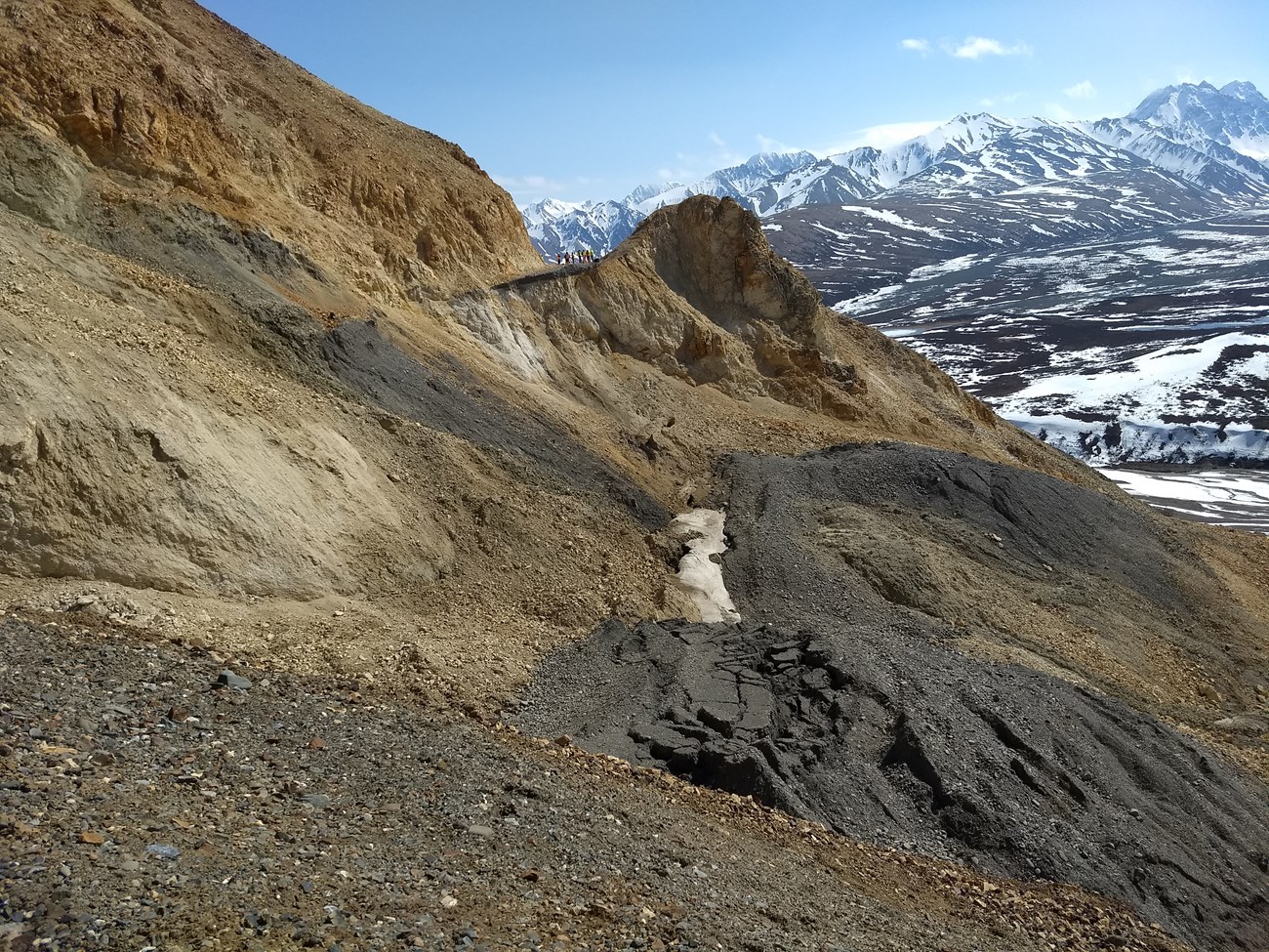 A rocky slope of gray, brown and orange rock on the side of a steep mountain slumps away downhill.