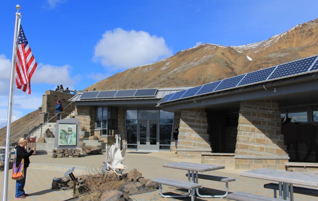 A stone building with big windows, solar panels on the roof and large mountain behind it.