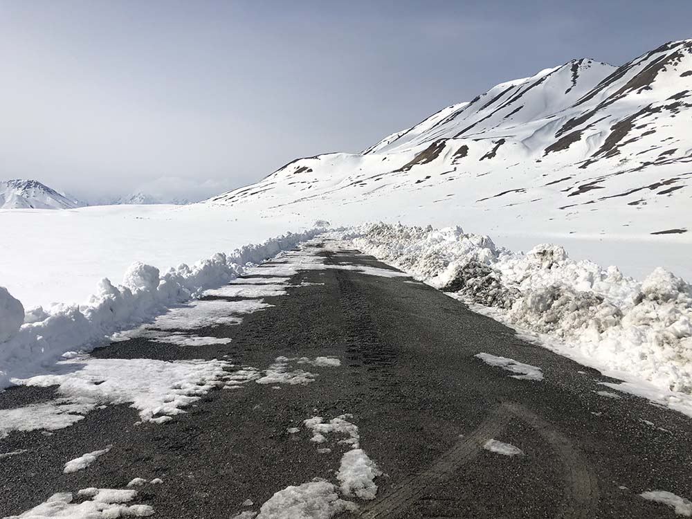 a snowy landscape of mountains and open areas. a dirt road is partially plowed in the foreground