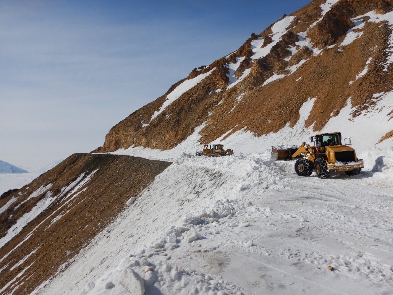 Two large plows clear snow from a narrow road set on the side of a steep mountain. Orange, red, and brown rocks make up the mountain where it's not covered by snow.