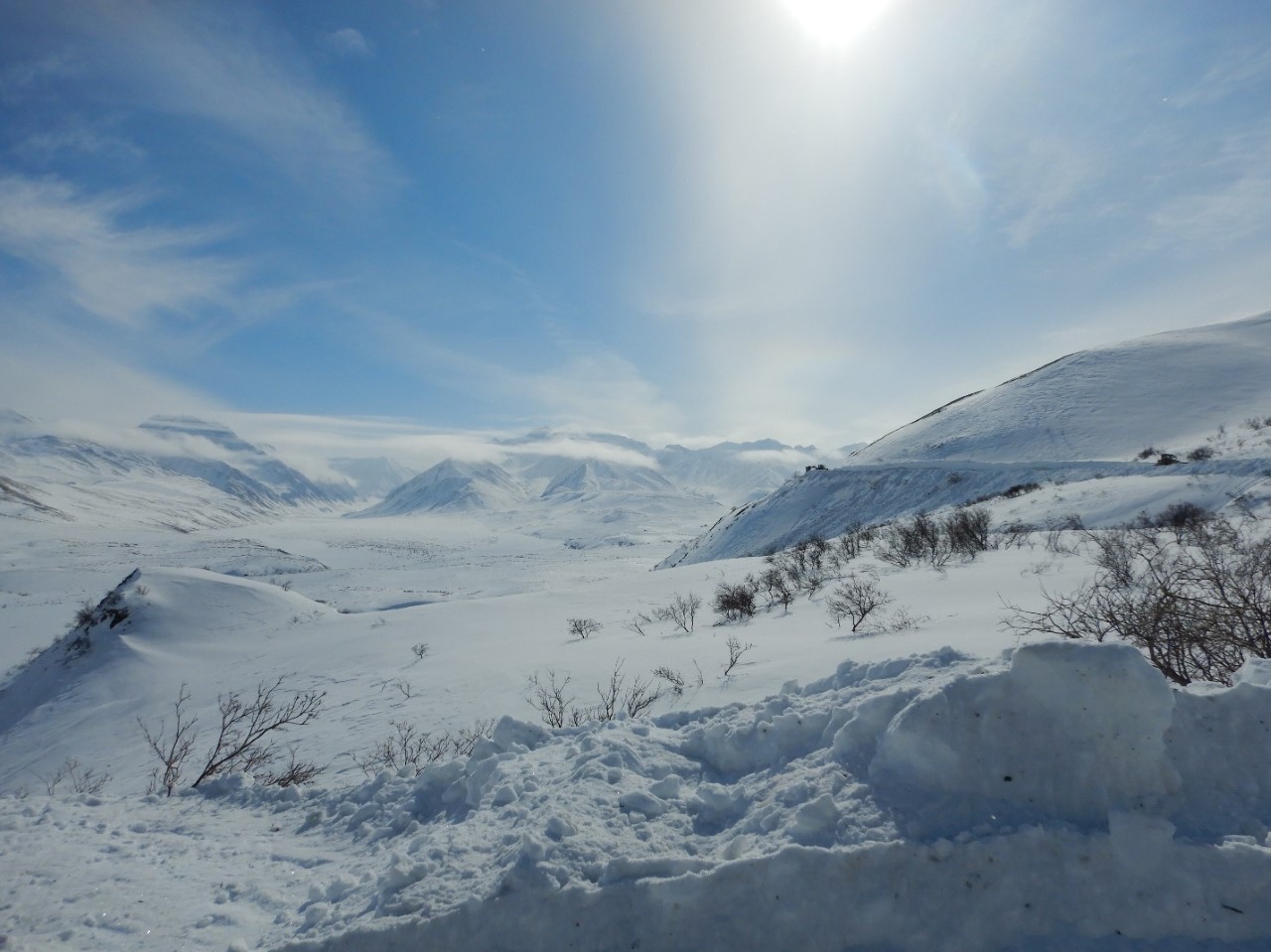 A snowy landscape of vast valleys and mountains. In the far distance, a tiny silhouette of a snowplow clearing the road can be seen.