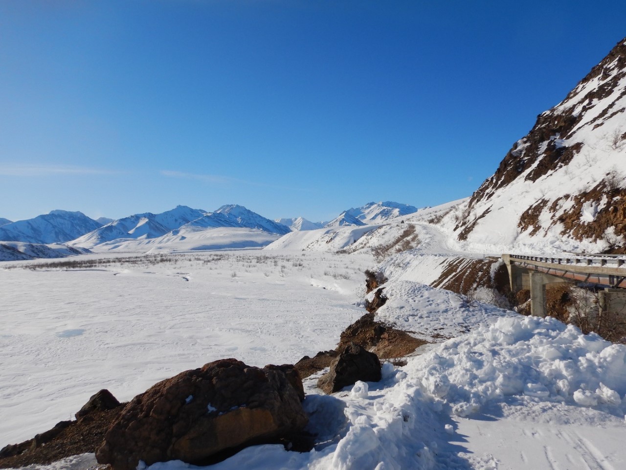 A snow-covered valley with mountains in the distance. On the right, a road crosses a bridge and then leads uphill. Plow berms are visible on the side of the road, though some snow still covers the road surface.