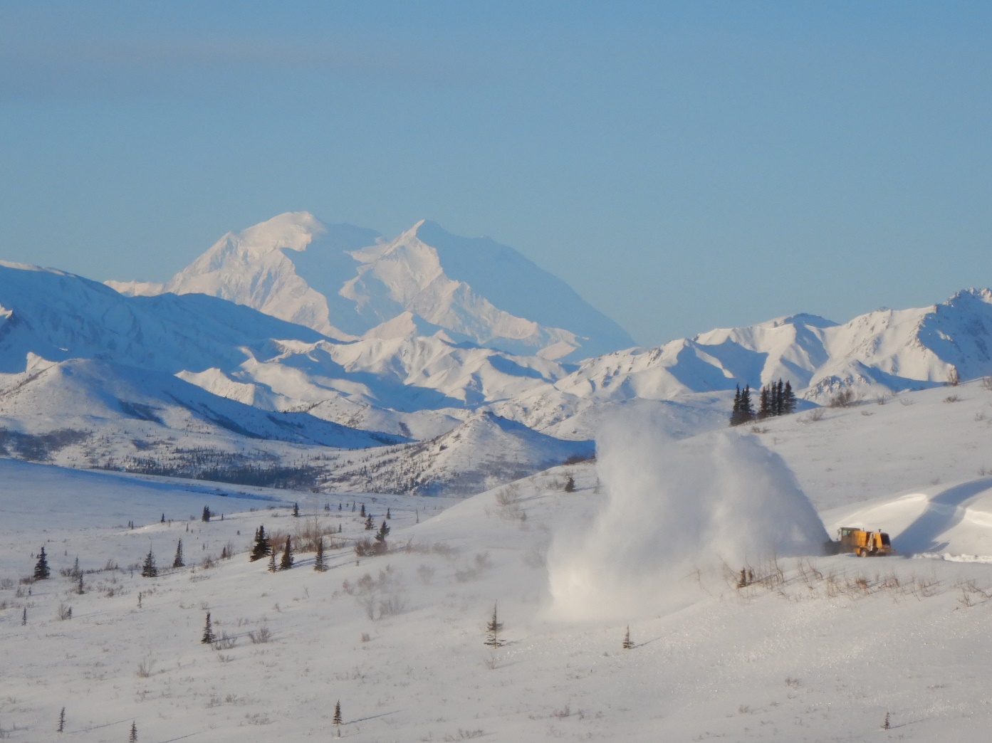 A wintry landscape on a sunny day with Denali looming large on the horizon. In the corner, a large orange snowblower clears the road and sends a large cloud of snow up into the air off to the side of the road.