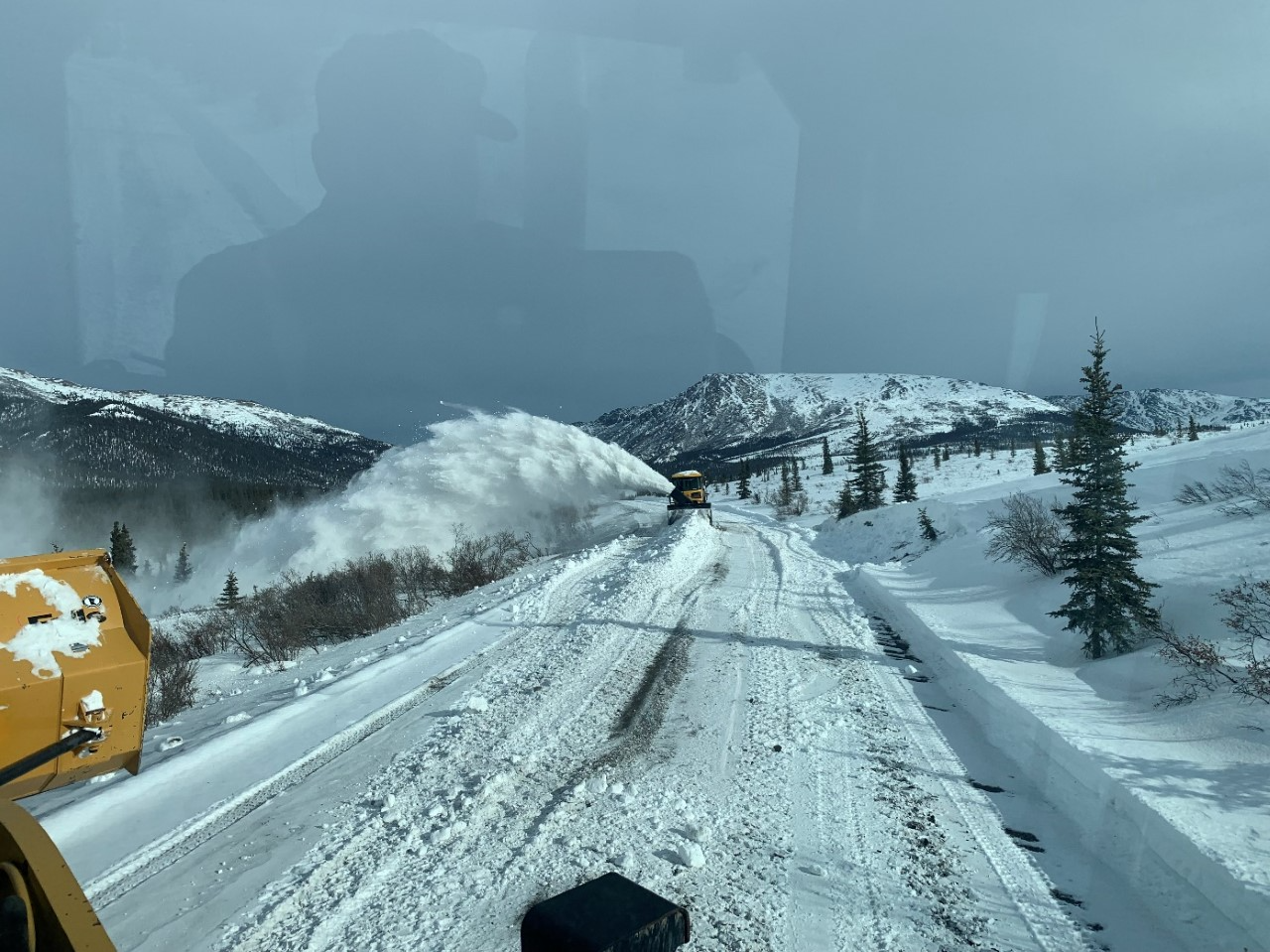 A snowblower clears snow from the road by sending it far off to the left side of the road in a high snow plume. A few inches of snow remains on the road, but the gravel surface of the road can just be seen through the snow in a few spots.