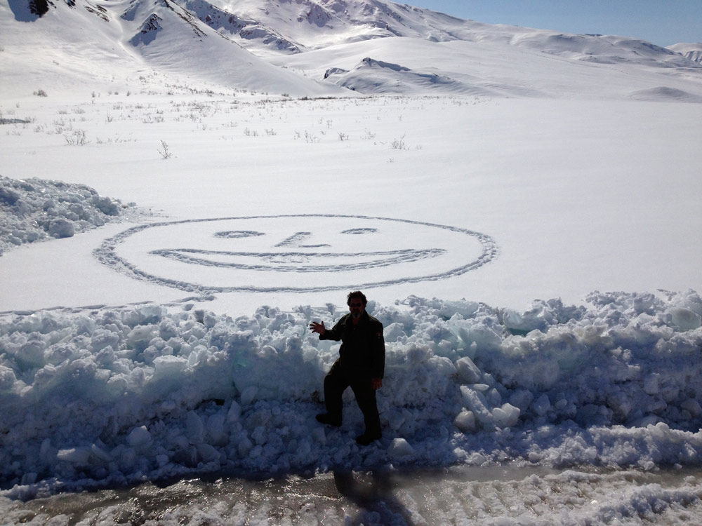 man standing near a snowy field in which a smiley face has been stomped in the snow