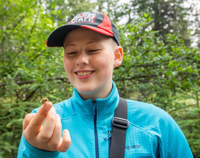 A student in a blue jacket and ball cap smiles at a bumble bee on his finger.