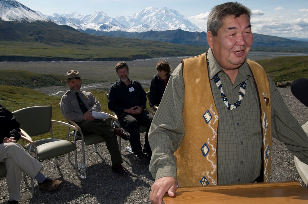 an alaska native man speaking at a lectern outside in front of several seated people. denali, a vast snowy mountain, is in the distance