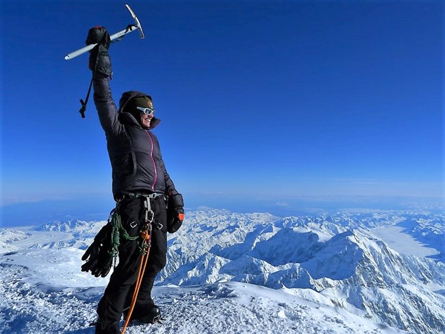 A mountaineer stands on the summit of Denali, overlooking a vast landscape of smaller, snowy mountains.
